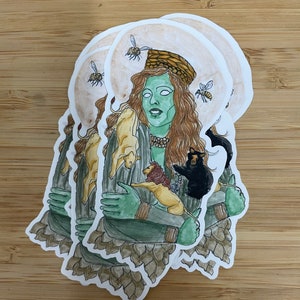 Puede incluir: Una pegatina con una mujer de piel verde con cabello largo y castaño que lleva un sombrero tejido. Está sosteniendo un león, un oso y dos animales más pequeños. El fondo es de color marrón claro con abejas volando alrededor de su cabeza.