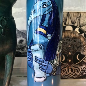 May include: A blue candle with a hand-painted image of a blue-skinned, bird-headed figure holding a scroll. The figure has a gold collar and blue hair.