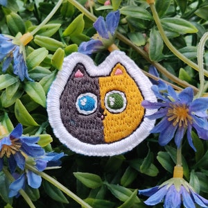 May include: A white, black, and yellow embroidered cat patch with a split face. One side is black with a blue eye and the other side is yellow with a green eye.