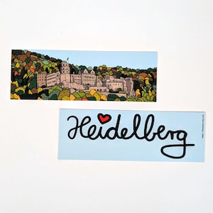 Könnte beinhalten: Ein blaues und weißes Lesezeichen mit einer schwarzen Illustration des Textes "Heidelberg" mit einem roten Herzen über dem Wort. Das Lesezeichen zeigt auch ein kleineres Bild einer Burg in einer Waldlandschaft.
