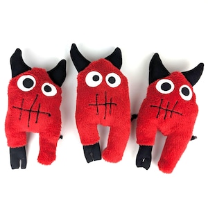 May include: Three red plush monster toys with black horns, eyes, and feet. Each has a stitched mouth and a fluffy texture. The toys are arranged on a white background.