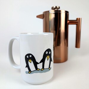 Könnte beinhalten: Ein weißer Keramikbecher mit einer Cartoon-Illustration von zwei Pinguinen, die sich auf einer hellblauen Eisfläche an den Händen halten, vor einer glänzenden kupferfarbenen French Press aus Metall mit zylindrischem Körper und Seitengriff.
