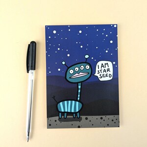 Könnte beinhalten: Ein blauer und grüner Comic-Alien mit fünf Augen und einer Sprechblase, die "I AM STAR SEED" sagt, vor einem dunkelblauen Hintergrund mit weißen Sternen.