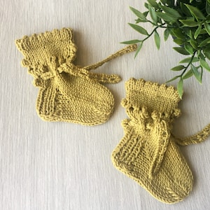 May include: Two mustard yellow knitted baby booties with a drawstring closure and a ruffled top edge. The booties are made of a soft, textured yarn and are displayed on a light-colored surface.