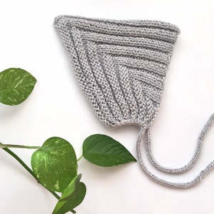Puede incluir: Un gorro de bebé de punto gris con una larga corbata. El gorro está hecho de un hilo suave y texturizado y tiene un diseño simple y clásico.
