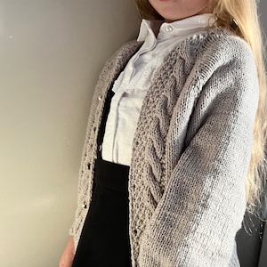 May include: A light gray cable knit cardigan with a white button-up shirt underneath. The cardigan has a cable knit pattern and is worn over a black skirt.