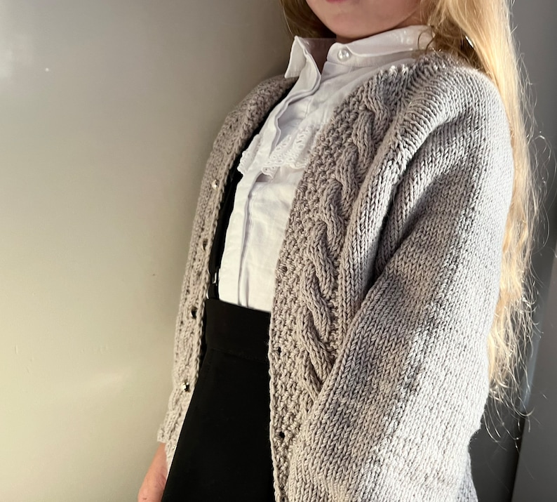 May include: A light gray cable knit cardigan with a white button-up shirt underneath. The cardigan has a cable knit pattern and is worn over a black skirt.