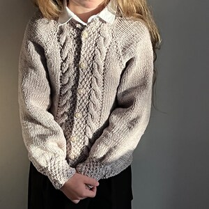 May include: A beige knitted cardigan with a cable knit pattern and pearl buttons. The cardigan has a round neckline and long sleeves.