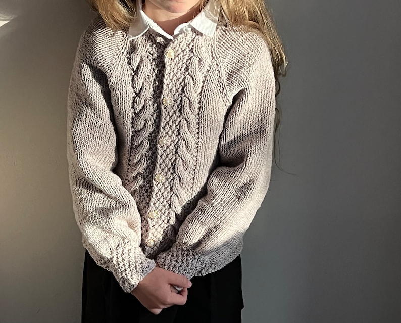 May include: A beige knitted cardigan with a cable knit pattern and pearl buttons. The cardigan has a round neckline and long sleeves.