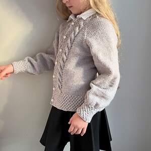 May include: A light gray knitted cardigan with a cable knit pattern and pearl buttons. The cardigan has long sleeves and a relaxed fit.