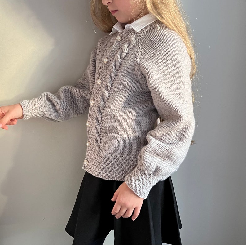 May include: A light gray knitted cardigan with a cable knit pattern and pearl buttons. The cardigan has long sleeves and a relaxed fit.