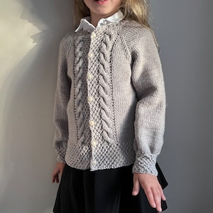 May include: A gray knitted cardigan with a cable knit pattern and button closure. The cardigan has a round neckline and long sleeves.
