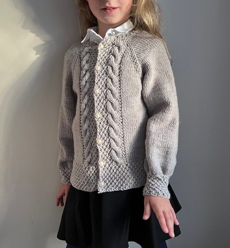 May include: A gray knitted cardigan with a cable knit pattern and button closure. The cardigan has a round neckline and long sleeves.