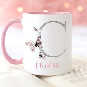 A white ceramic mug with a pink rim and handle. The mug has a silver letter 'C' with butterflies on the side. The mug has the name 'Charlotte' written in pink below the letter.