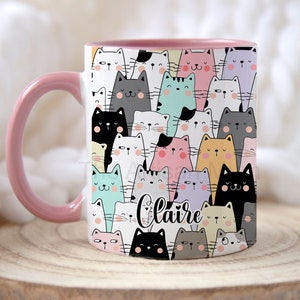 May include: White ceramic mug with a pink handle and interior. The mug features a pattern of cartoon cats in various colours, including black, grey, pink, and blue. The name "Claire" is written in cursive on the front.