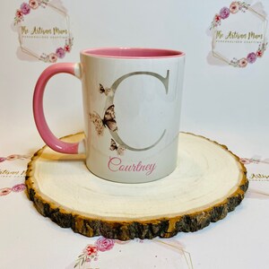 Puede incluir: Una taza de cer&aacute;mica blanca con un borde y un asa de color rosa. La taza tiene una letra "C" gris y un dise&ntilde;o de mariposa. El nombre "Courtney" est&aacute; escrito debajo de la letra. La taza est&aacute; sobre una rodaja de madera.