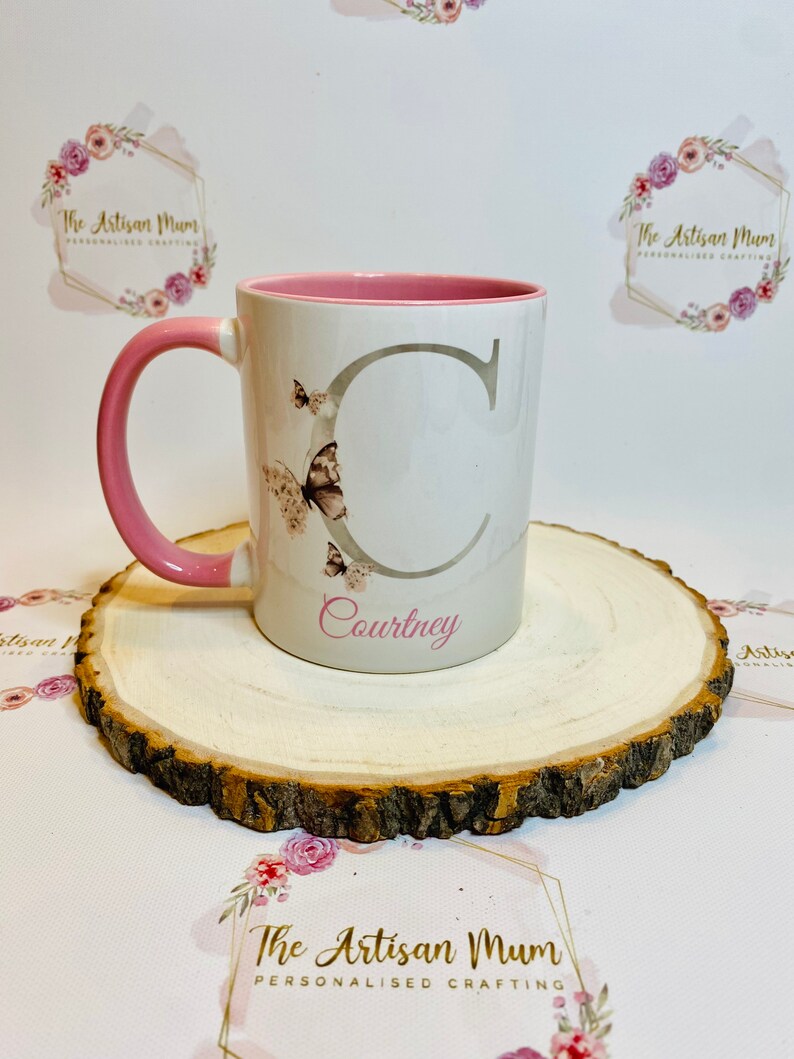 Puede incluir: Una taza de cer&aacute;mica blanca con un borde y un asa de color rosa. La taza tiene una letra "C" gris y un dise&ntilde;o de mariposa. El nombre "Courtney" est&aacute; escrito debajo de la letra. La taza est&aacute; sobre una rodaja de madera.