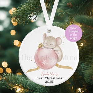 Può includere: Ornamento natalizio rotondo con un'illustrazione di un topo che dorme su una palla rosa. L'ornamento ha il testo "Isabella's First Christmas 2025" e un nastro bianco per appenderlo.