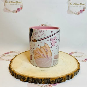 Pu&ograve; includere: Una tazza in ceramica bianca con un bordo rosa e un design con una mano con smalto rosa, un pennello e cuori. La tazza &egrave; su un sottobicchiere di legno.