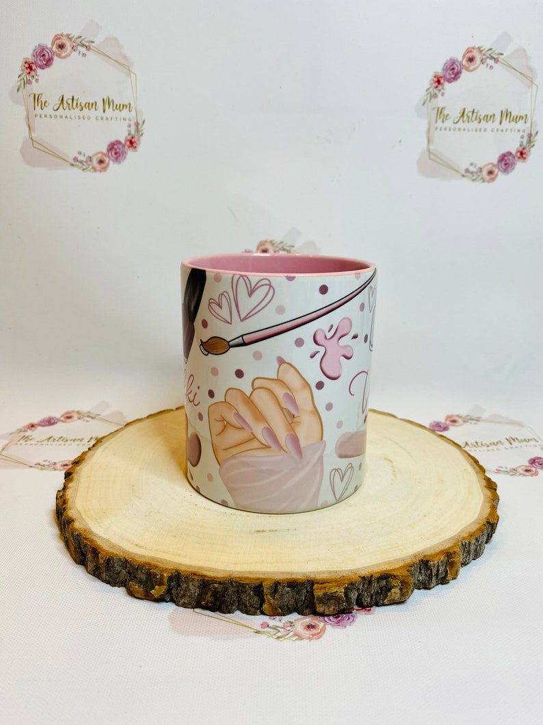 Pu&ograve; includere: Una tazza in ceramica bianca con un bordo rosa e un design con una mano con smalto rosa, un pennello e cuori. La tazza &egrave; su un sottobicchiere di legno.