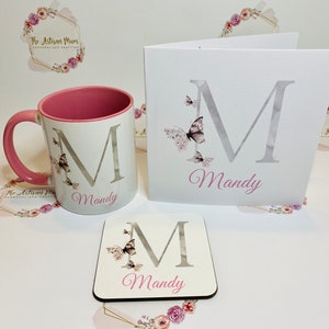 Puede incluir: Un conjunto de regalo personalizado que incluye una taza blanca con borde rosa, un posavasos y una tarjeta. Todos los art&iacute;culos presentan una letra "M" plateada con mariposas y el nombre "Mandy" en letras rosas.