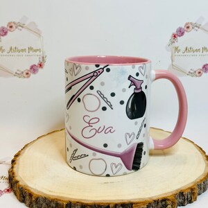 Peut inclure: Une tasse en c&eacute;ramique rose et blanche avec une anse rose. La tasse est d&eacute;cor&eacute;e d'outils de coiffure, notamment des ciseaux, une brosse &agrave; cheveux, un flacon pulv&eacute;risateur et des pinces &agrave; cheveux. Le nom "Eva" est &eacute;crit en rose sur la tasse.