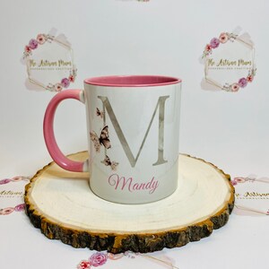Puede incluir: Una taza de cer&aacute;mica blanca con un borde y un asa de color rosa. La taza presenta una gran letra "M" plateada con un efecto acuarela y un dise&ntilde;o de mariposa. El nombre "Mandy" est&aacute; escrito en rosa debajo de la letra. La taza est&aacute; sentada sobre una rodaja de madera.