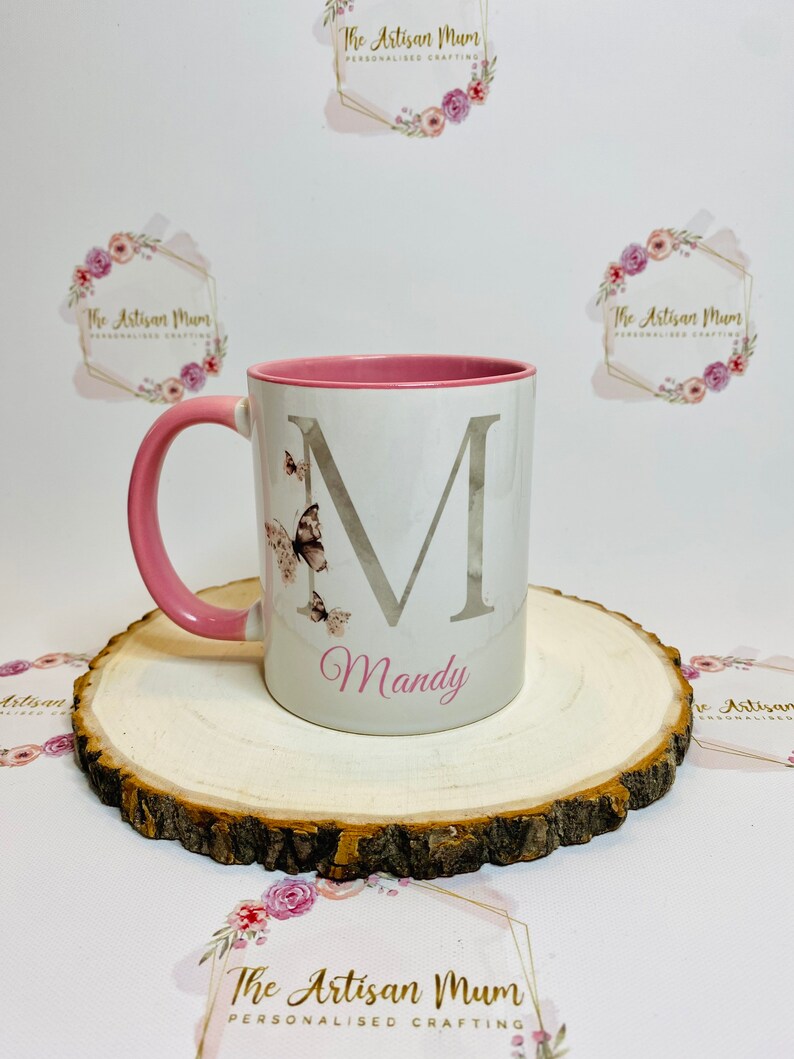 Puede incluir: Una taza de cer&aacute;mica blanca con un borde y un asa de color rosa. La taza presenta una gran letra "M" plateada con un efecto acuarela y un dise&ntilde;o de mariposa. El nombre "Mandy" est&aacute; escrito en rosa debajo de la letra. La taza est&aacute; sentada sobre una rodaja de madera.