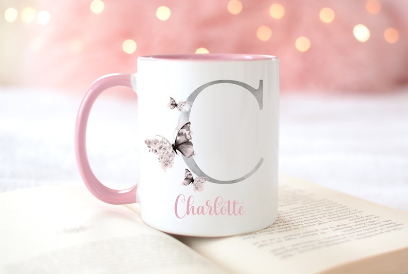 A white ceramic mug with a pink rim and handle. The mug has a silver letter 'C' with butterflies on the side. The mug has the name 'Charlotte' written in pink below the letter.