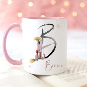 Pu&ograve; includere: Una tazza in ceramica bianca con un bordo e un manico rosa. La tazza presenta un'illustrazione in bianco e nero della lettera "B" con pennelli per il trucco e rossetto all'interno della lettera. Il nome "Bonnie" &egrave; scritto in corsivo rosa sotto la lettera.