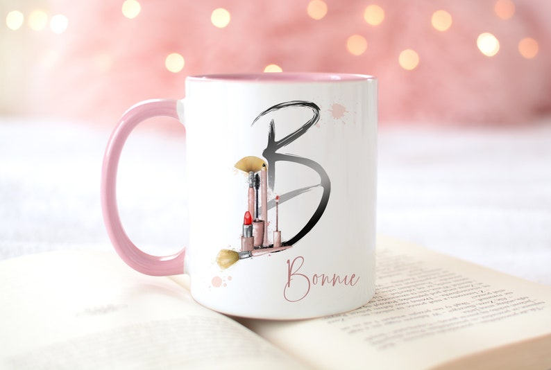 Pu&ograve; includere: Una tazza in ceramica bianca con un bordo e un manico rosa. La tazza presenta un'illustrazione in bianco e nero della lettera "B" con pennelli per il trucco e rossetto all'interno della lettera. Il nome "Bonnie" &egrave; scritto in corsivo rosa sotto la lettera.