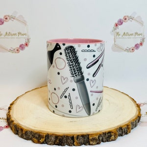 Peut inclure: Une tasse en c&eacute;ramique blanche avec un bord rose pr&eacute;sente un motif d'outils de coiffure, notamment un peigne, des ciseaux et des pinces &agrave; cheveux. Le motif est en noir et gris sur un fond blanc avec des accents roses.
