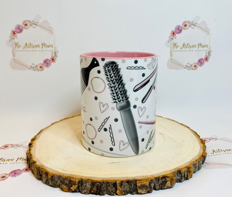 Peut inclure: Une tasse en c&eacute;ramique blanche avec un bord rose pr&eacute;sente un motif d'outils de coiffure, notamment un peigne, des ciseaux et des pinces &agrave; cheveux. Le motif est en noir et gris sur un fond blanc avec des accents roses.