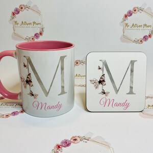Puede incluir: Una taza blanca con un borde y un asa de color rosa, con una letra "M" plateada con detalles de mariposa y el nombre "Mandy" impreso debajo. Tambi&eacute;n se muestra un posavasos a juego con el mismo dise&ntilde;o.