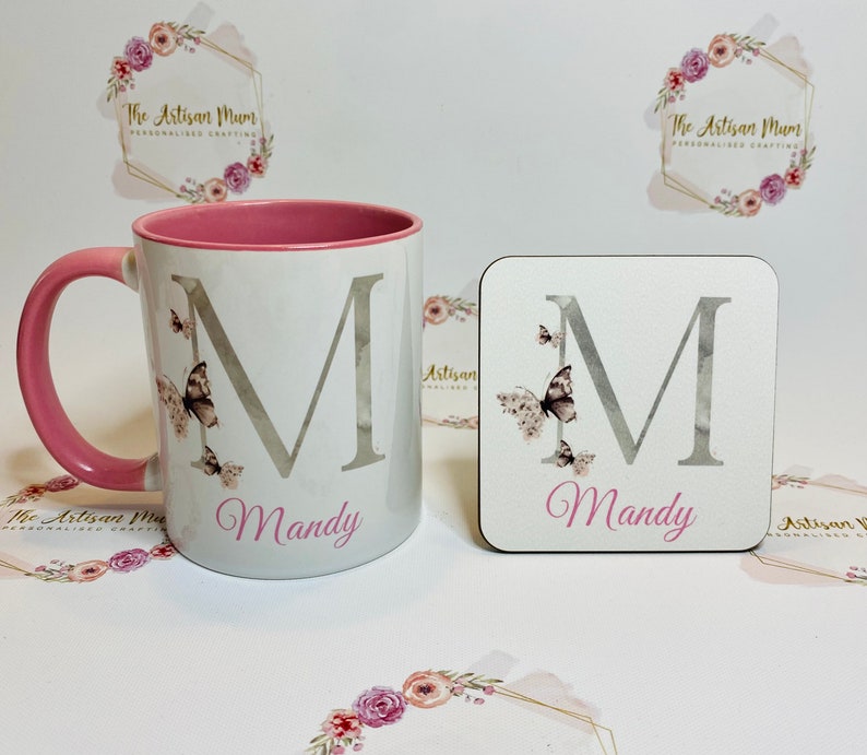 Puede incluir: Una taza blanca con un borde y un asa de color rosa, con una letra "M" plateada con detalles de mariposa y el nombre "Mandy" impreso debajo. Tambi&eacute;n se muestra un posavasos a juego con el mismo dise&ntilde;o.