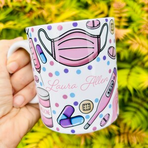 May include: White ceramic mug with a pink face mask, pills, a thermometer, and the name "Laura Allen" printed on it. The mug is decorated with pink, blue, and purple polka dots.