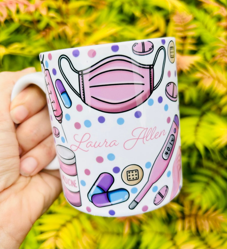 May include: White ceramic mug with a pink face mask, pills, a thermometer, and the name "Laura Allen" printed on it. The mug is decorated with pink, blue, and purple polka dots.