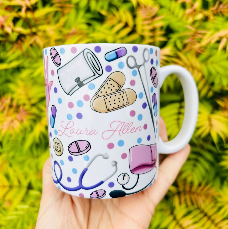 May include: White ceramic mug with a colorful pattern of medical supplies, including bandages, pills, a stethoscope, and a pair of scissors. The mug has the name "Laura Allen" written in pink cursive on the front.