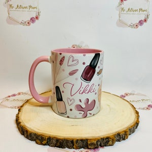 Pu&ograve; includere: Una tazza in ceramica rosa e bianca con un manico rosa. La tazza ha un design di bottiglie di smalto per unghie, cuori e schizzi di vernice rosa. Il nome "Vikki" &egrave; scritto in un font rosa su la tazza.