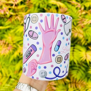 May include: White ceramic mug with a pink and blue design featuring medical symbols like pills, a stethoscope, a thermometer, and a gloved hand. The mug has the name "Lauren" written on it.