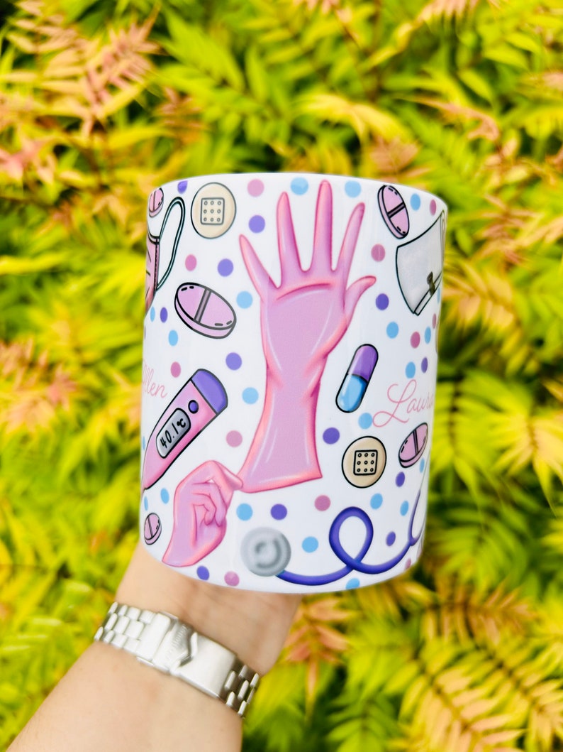 May include: White ceramic mug with a pink and blue design featuring medical symbols like pills, a stethoscope, a thermometer, and a gloved hand. The mug has the name "Lauren" written on it.