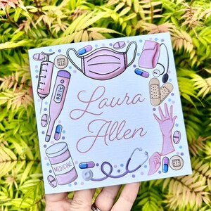 May include: A blue and white card with a pink medical theme. The card has a pink face mask, a thermometer, a syringe, a stethoscope, a bottle of medicine, a pair of gloves, and band-aids. The card says "Laura Allen" in pink cursive writing.