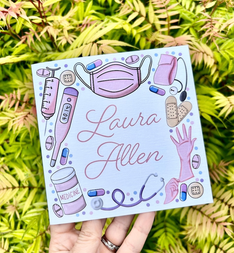 May include: A blue and white card with a pink medical theme. The card has a pink face mask, a thermometer, a syringe, a stethoscope, a bottle of medicine, a pair of gloves, and band-aids. The card says "Laura Allen" in pink cursive writing.