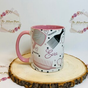 Peut inclure: Une tasse en c&eacute;ramique rose et blanche avec une anse rose. La tasse est d&eacute;cor&eacute;e de peignes &agrave; cheveux, d'un miroir et du nom "Eva" en lettres roses.