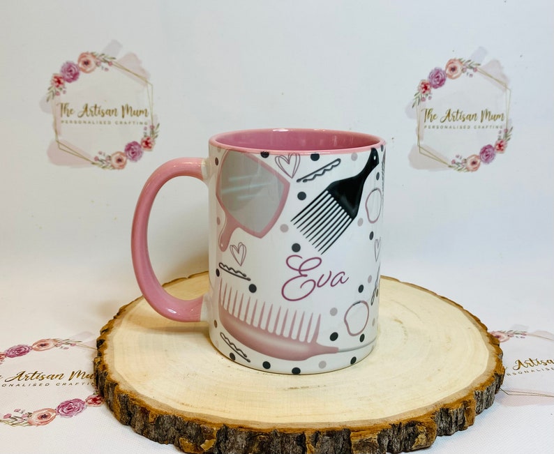 Peut inclure: Une tasse en c&eacute;ramique rose et blanche avec une anse rose. La tasse est d&eacute;cor&eacute;e de peignes &agrave; cheveux, d'un miroir et du nom "Eva" en lettres roses.