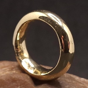 May include: A solid gold ring with a polished finish. The ring is a simple band design, with a smooth, rounded surface. The ring is displayed on a brown, textured surface, with a dark background.