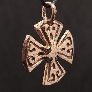 Può includere: Un ciondolo color bronzo a forma di croce, sospeso a un cordino nero. La croce presenta un design intricato e strutturato con motivi geometrici e dettagli circolari. Il ciondolo ha un aspetto leggermente invecchiato.