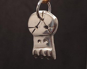 May include: A silver pendant shaped like a skull with a small loop at the top for hanging. The skull has a detailed design with a cross-hatched pattern on the top and two small circles on the bottom.