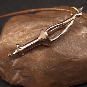 May include: A bronze-coloured pendant necklace with an abstract design. The pendant hangs from a thin, brown cord. The necklace is displayed on a textured, light brown stone surface.