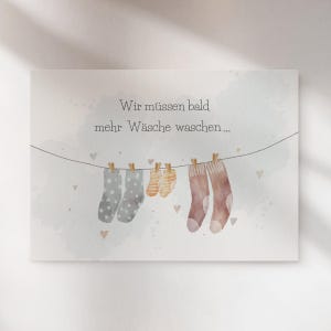 May include: A rectangular greeting card featuring a watercolour illustration of a clothesline with two grey polka-dot socks, two small yellow striped baby socks, and two brown socks hanging from wooden clips, with the text Wir m&uuml;ssen bald mehr W&auml;sche waschen.